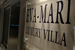 En skylt med texten ”EVA-MARI LUXURY VILLA”, med en metallisk yta och tydliga, framträdande bokstäver.