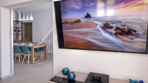 A modern living room with a large flat-screen TV displaying a beach scene, a dining area with a wooden table and blue chairs, and a small console table with a game controller and decorative items. - Villa Eva Mari - Villa Eva Mari (Chania)