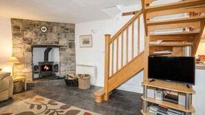 A living room with a stone fireplace, a wooden staircase, and a flat-screen TV on a wooden stand. - Beech Tree Cottage (Caernarfon)