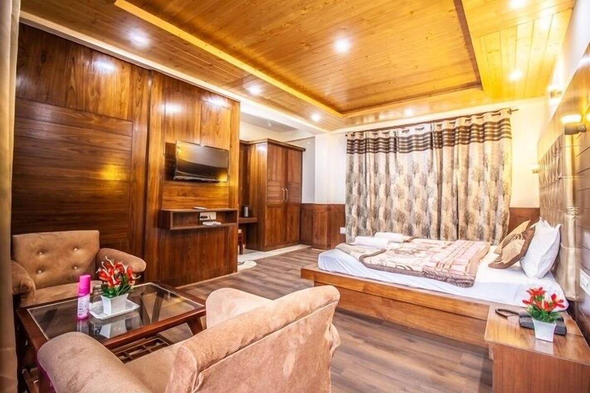 A room with a wooden ceiling, a bed with a patterned bedspread, a television mounted on the wall, a sofa, and a coffee table with a vase of flowers.