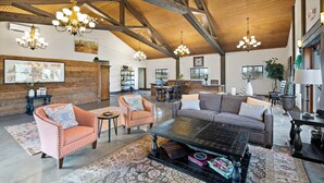 Un vaste salon doté d’un haut plafond, de poutres en bois et de plusieurs lustres. On y trouve des coins salon confortables avec des canapés et des fauteuils, ainsi qu’une table basse au centre.
