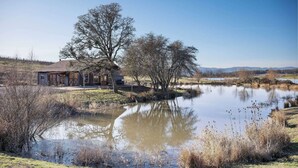 Une maison rustique au bord d’un étang, sous un ciel dégagé.