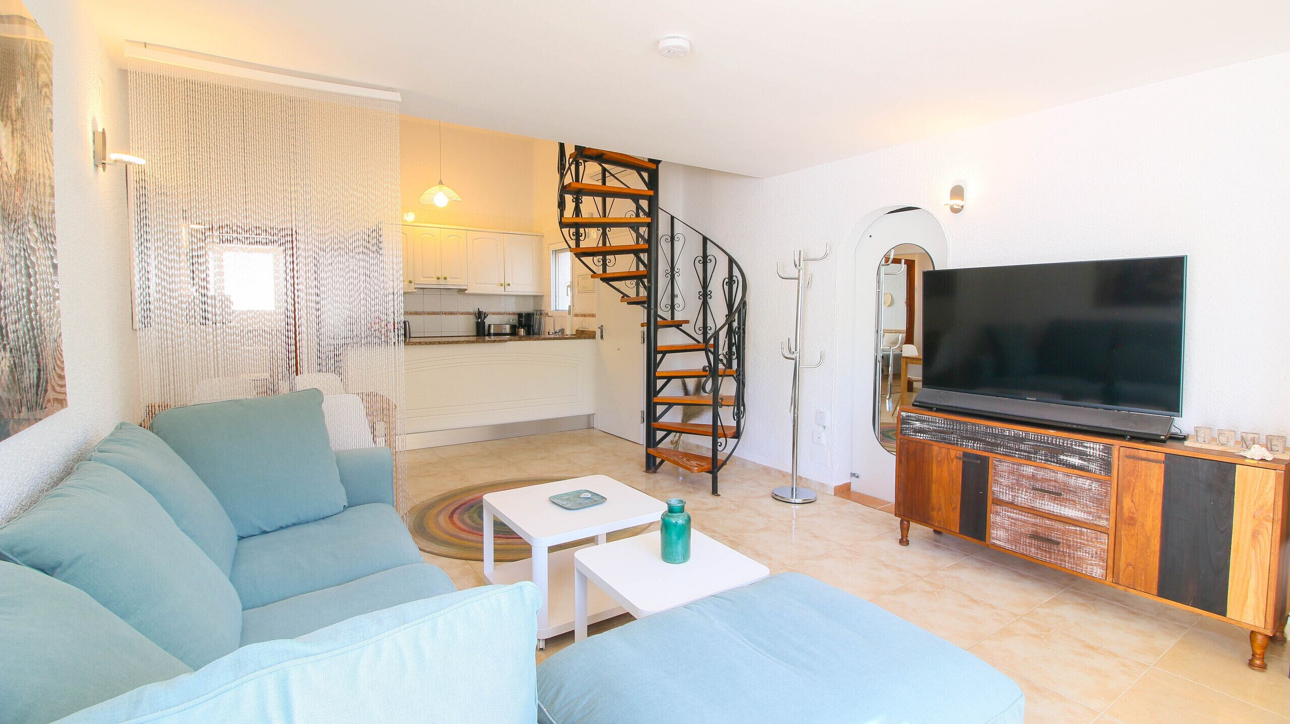 Una sala de estar con un sofá, una mesa de centro, un televisor, una escalera y una cocina al fondo.