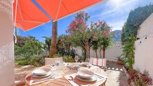 A round wooden table set for two with white plates, glasses, and cutlery, under an orange umbrella, surrounded by potted plants and a white wall. - Nice Bungalow next to Denia (Denia)