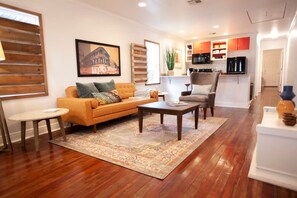 A living room with a sofa, coffee table, and a rug. There is a kitchen in the background with cabinets and a refrigerator.