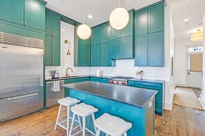 A modern kitchen with teal cabinets, a stainless steel refrigerator, a central island with stools, and pendant lighting. - Family Perfect Point Home |  Luxury at its Best (New Orleans)