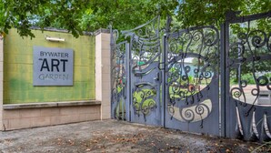 Una reja metálica con diseños intrincados, un muro verde con un cartel que dice “BYWATER ART GARDEN” y un sendero pavimentado con hojas dispersas.