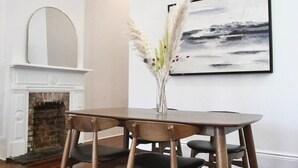 A dining area with a wooden table, four chairs, a vase with pampas grass, a fireplace, and a framed abstract painting on the wall.