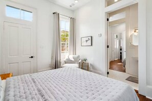 A bedroom with a large bed, a window with curtains, a bedside table with a plant, and a door leading to another room.