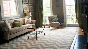 Ein Wohnzimmer mit einer Couch, einem Sessel und einem Couchtisch, großen Fenstern mit Vorhängen sowie Wandbildern.