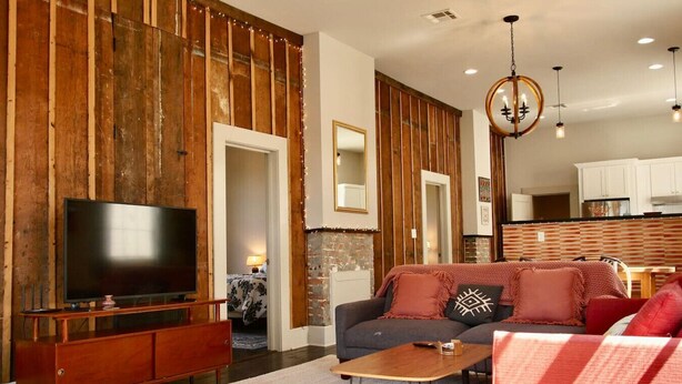 A living room with a TV, sofa, coffee table, wooden wall paneling, and a kitchen in the background.