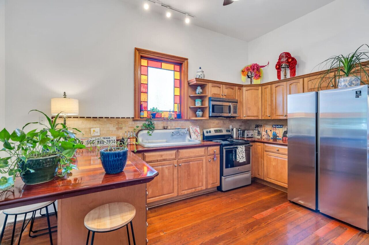 A kitchen with wooden cabinets, a stainless steel refrigerator, a microwave, and a stove. There are plants on the counter and a window with a stained glass design.