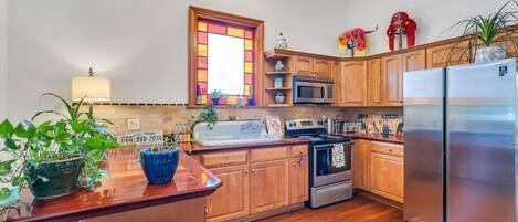 A kitchen with wooden cabinets, a stainless steel refrigerator, a microwave, and a stove. There are plants on the counter and a window with a stained glass design.