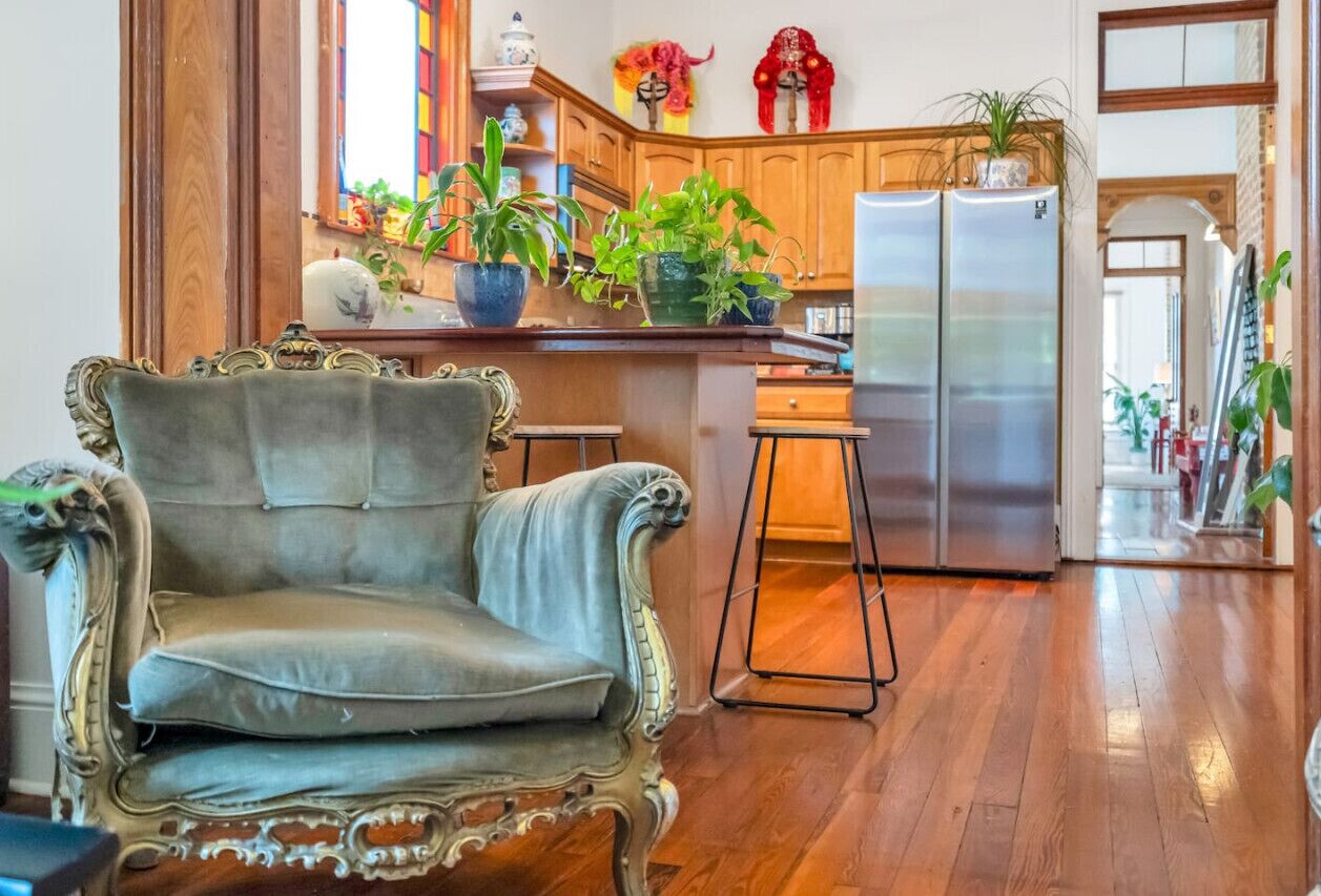 Een ruime kamer met een grote, versierde stoel, een houten vloer en een keuken met houten kastjes en een roestvrijstalen koelkast.