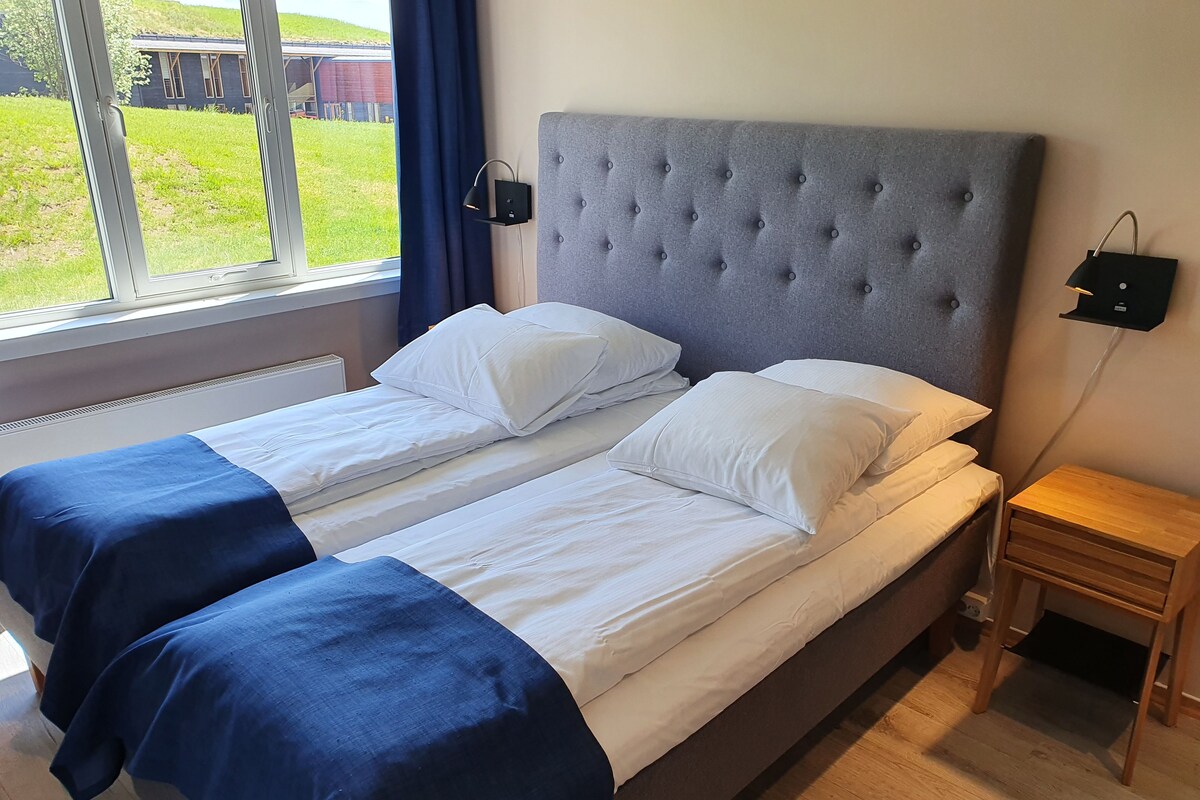 Una cama doble con cabecera tapizada en gris, ropa de cama blanca y un detalle azul en el piecero. Hay una mesita de noche con una lámpara y un enchufe en la pared.