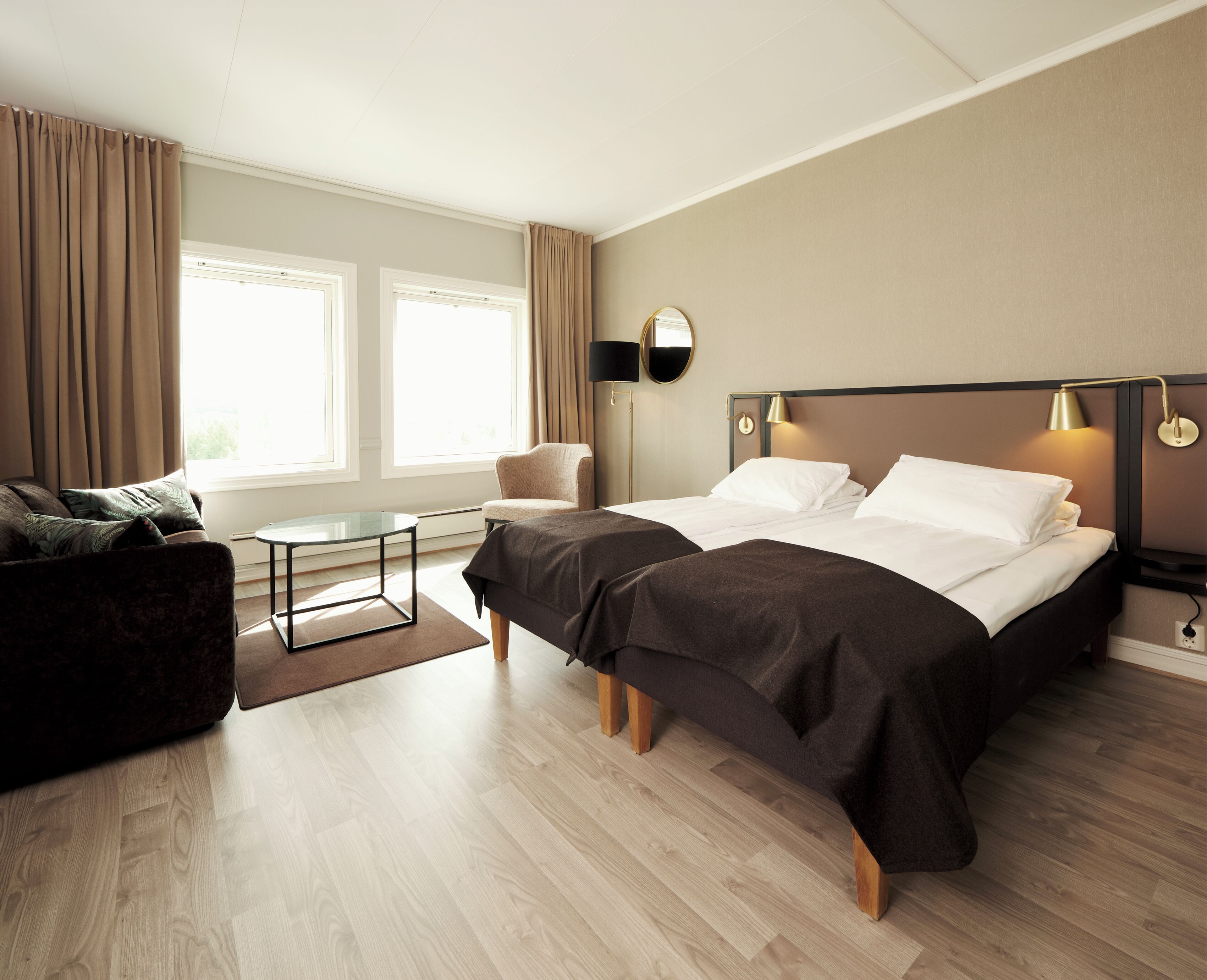 A hotel room with a bed, a sofa, a small table, and a chair. There are two windows with curtains, a lamp on the wall, and a mirror.