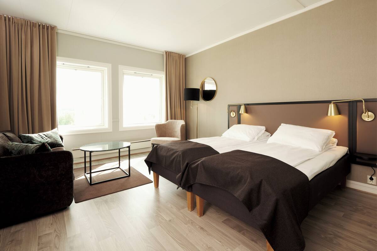 A hotel room with a bed, a sofa, a small table, and a chair. There are two windows with curtains, a lamp on the wall, and a mirror.