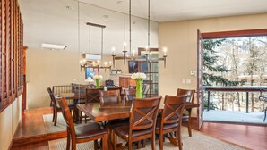 A dining area with a large wooden table and chairs, a chandelier, and a view of trees through a glass door.