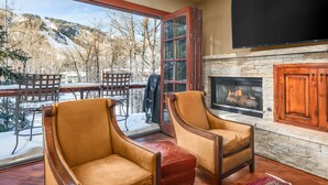A cozy room with a fireplace, two leather armchairs, a dining table with chairs, and a large window overlooking a snowy landscape.