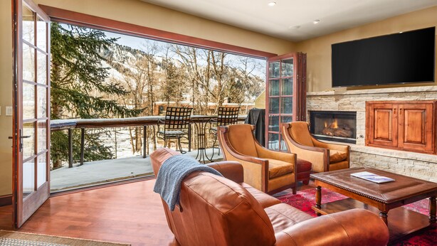 A spacious living room with leather sofas, a fireplace, a flat-screen TV, and a large sliding glass door leading to a balcony with a view of trees and a mountain.