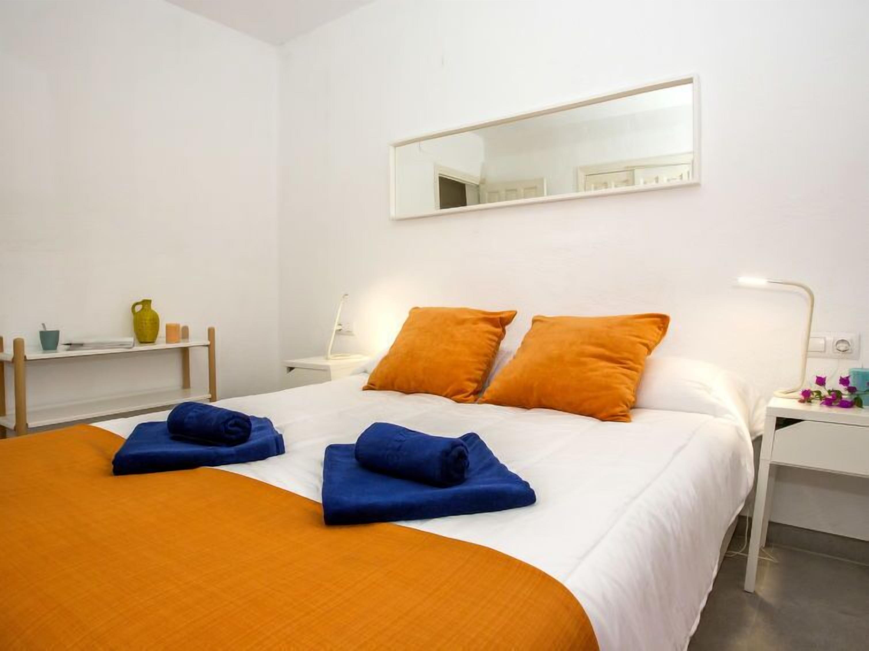 Una habitación de hotel con una cama, dos almohadas naranjas, una mesita de noche blanca, un espejo y una estantería con un florero y velas.