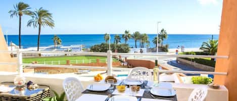 Un balcón con mesa lista para dos personas, con vista a una playa con palmeras y un cielo azul despejado.