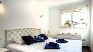 A neatly made bed with white bedding and blue pillows, a bedside table, a small desk with a lamp, and a window with a view of the outside. - Deltamar by Interhome (Jávea)