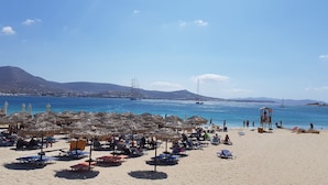 A beach with umbrellas and lounge chairs, a lifeguard tower, and boats in the water. - Parian Beach House with Pool Access, walk to the Beach (Paros)