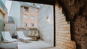 Une terrasse en bois avec des chaises longues et une table, un mur en pierre et une vue sur un immeuble doté de nombreuses fenêtres.