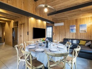 Een houten interieur met een eettafel gedekt voor vier personen, omringd door rieten stoelen. Een flatscreen televisie is aan de muur bevestigd, en een bank is op de achtergrond te zien.