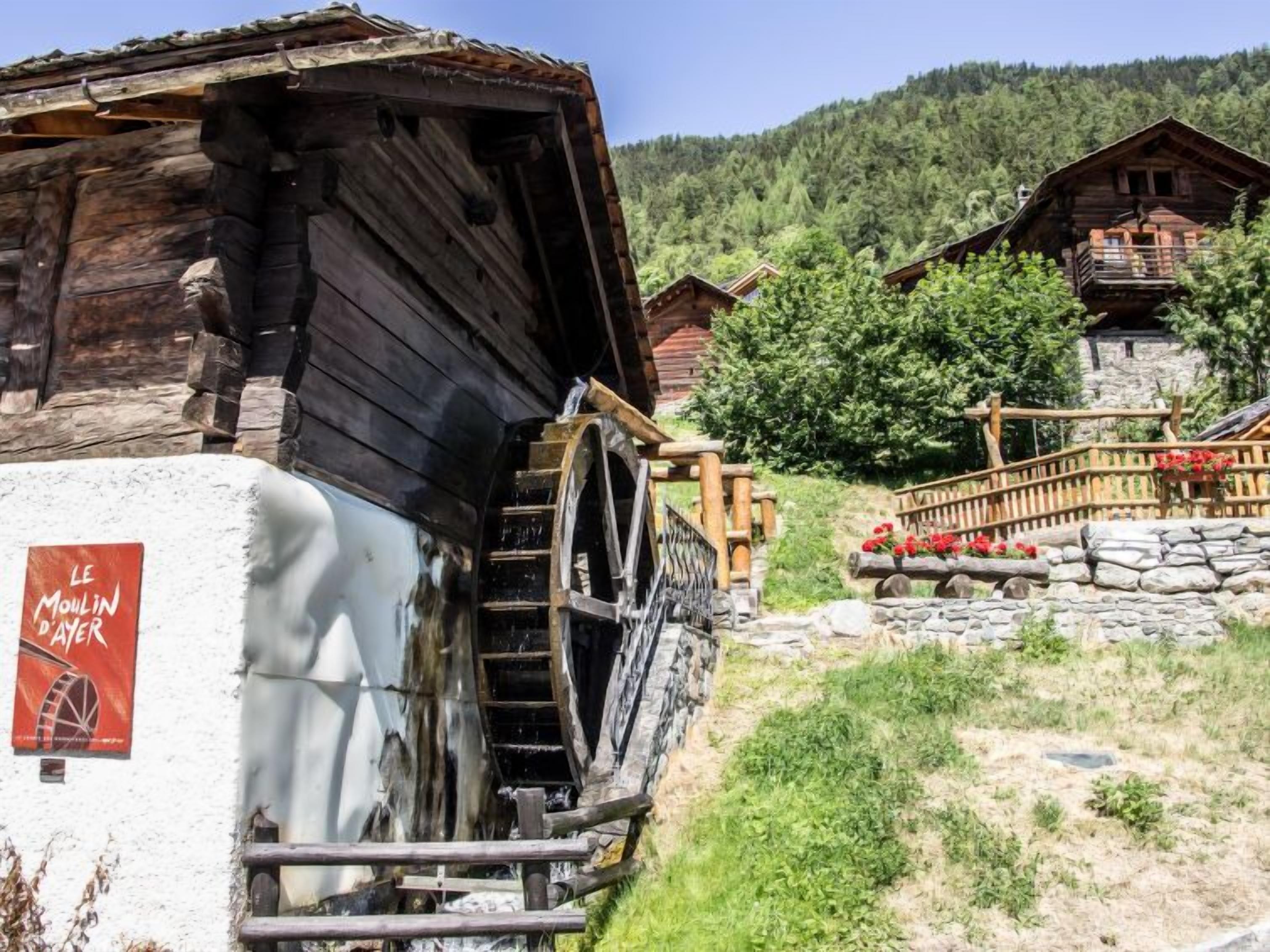 A rustic wooden building with a water wheel, surrounded by lush greenery and other wooden structures.