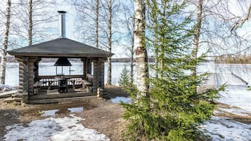 Puinen huvimaja, jossa on savupiippu, ja jonka ympärillä on koivuja ja maata, joka on peittynyt lumeen.