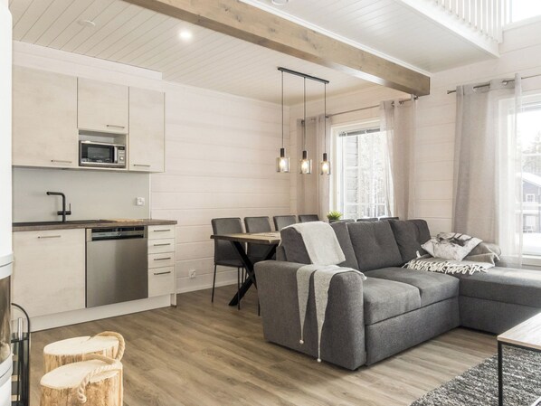 A modern kitchen with a microwave, dishwasher, and wooden cabinets. There is a dining area with a table and chairs, and a grey sofa in the living room.