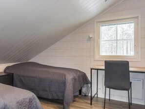 Un pequeño dormitorio en ático, con una sola habitación, techo inclinado, ventana con vistas a árboles, cama, escritorio y silla.