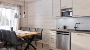 A modern kitchen with a wooden dining table, grey chairs, stainless steel appliances, and a window with curtains.