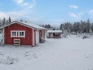 Une cabane en bois rouge, avec un toit enneigé et une petite construction adjacente, entourée d’un paysage hivernal et d’arbres.