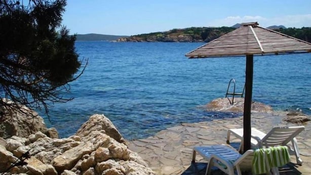 A stone patio with two lounge chairs and a small table, overlooking a body of water with a rocky shoreline and a distant hill. - La Nina by Interhome (Porto Rotondo)