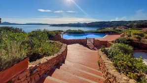 A pool area with a stone pathway leading to a pool, surrounded by greenery and overlooking a body of water with distant hills. - La Nina by Interhome (Porto Rotondo)
