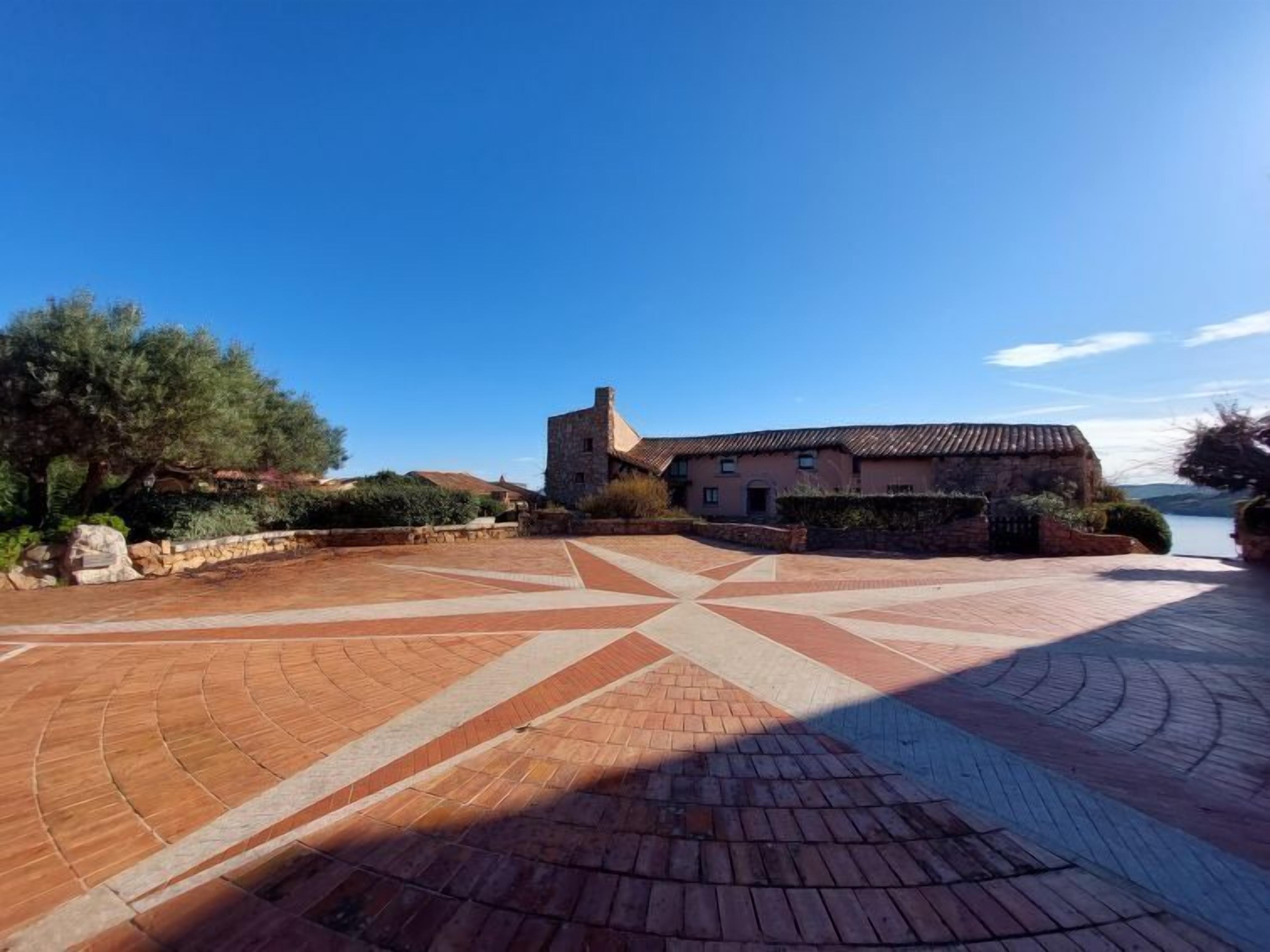A courtyard with a stone building, a paved area with geometric patterns, and a tree.