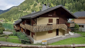 Un bâtiment de style chalet doté d’un balcon, situé près d’un lac et entouré de collines boisées.