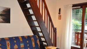 Une pièce avec un escalier en bois, un canapé bleu à motifs, un petit tabouret en bois et une porte-fenêtre coulissante donnant sur un espace extérieur.
