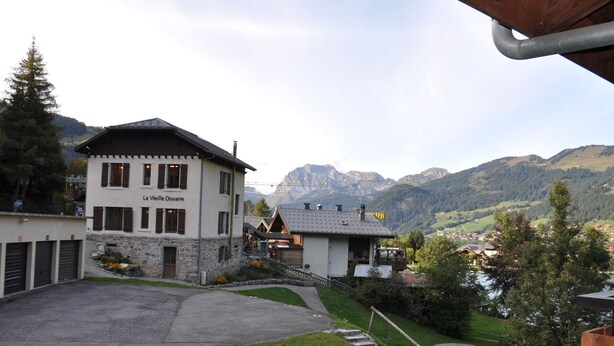 Un bâtiment appelé « La Vielle Douane », doté d’une façade en pierre, d’un escalier en bois menant à l’entrée du deuxième étage, et offrant une vue sur les montagnes en arrière-plan.