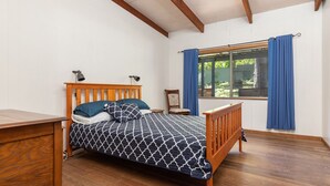 Ein Schlafzimmer mit einem hölzernen Bett, einem Nachttisch, einem Stuhl, einem Fenster mit blauen Vorhängen und Blick auf viel Grün.
