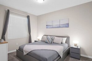 A bedroom with a bed, bedside tables, a nightstand, a window with curtains, and a painting on the wall.