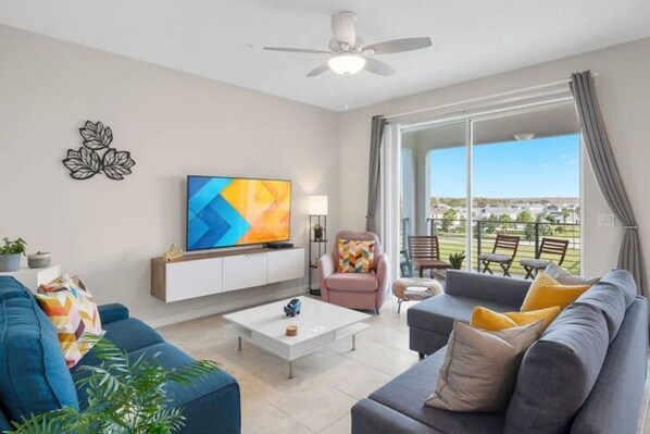 A modern living room with a flat-screen TV, a sofa, a coffee table, and a chair. There is a balcony with outdoor seating visible through large windows.