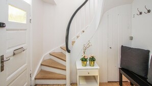 Una escalera con escalones de madera y una pared blanca. Hay una pequeña mesa blanca con dos macetas doradas de flores. Se ve un banco a la derecha.
