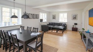Una sala moderna con un sofá oscuro, una mesa de centro y un comedor con una mesa y sillas oscuras. Hay cuadros enmarcados en las paredes y un ventanal que proporciona luz natural.
