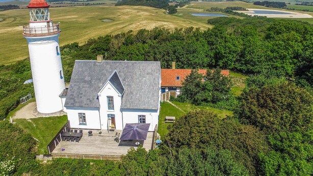 Un faro y un edificio blanco con techo gris, rodeado de vegetación y campos.