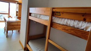 Un lit superposé avec un matelas, une table en bois et une chaise, dans une pièce dotée d’une fenêtre.