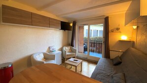 A compact living space with a sofa, two chairs, a small table, a TV mounted on the wall, and a sliding glass door leading to an outdoor area. - Cozy studio near slopes with private parking (Châtel)